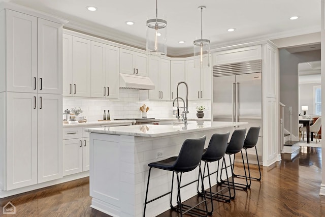 kitchen featuring hanging light fixtures, a center island with sink, built in fridge, custom range hood, and white cabinets