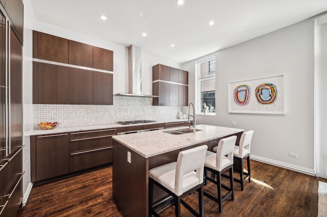 kitchen with wall chimney range hood, sink, a kitchen island with sink, backsplash, and a kitchen breakfast bar