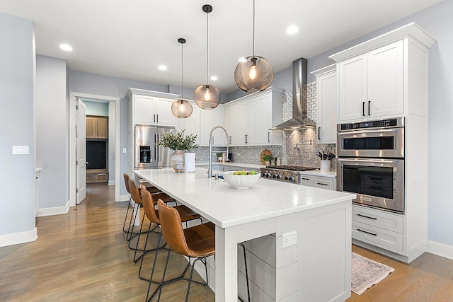 kitchen with a kitchen island with sink, pendant lighting, stainless steel appliances, and wall chimney exhaust hood