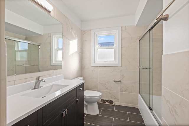 full bathroom featuring bath / shower combo with glass door, tile walls, vanity, and toilet