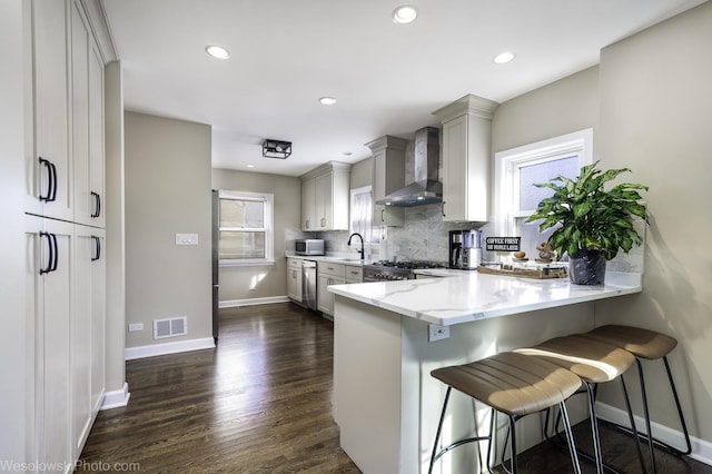 kitchen featuring wall chimney exhaust hood, a breakfast bar area, appliances with stainless steel finishes, kitchen peninsula, and decorative backsplash