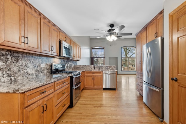 kitchen with light wood-type flooring, appliances with stainless steel finishes, ceiling fan, light stone countertops, and decorative backsplash