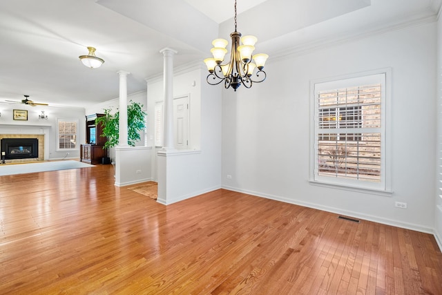 unfurnished dining area with ornate columns, ceiling fan with notable chandelier, a tiled fireplace, crown molding, and light wood-type flooring