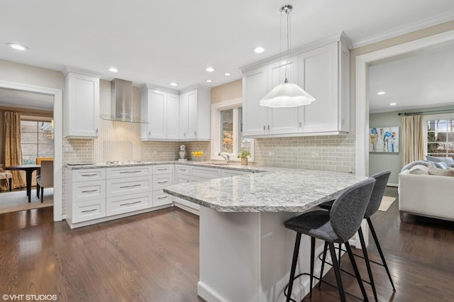 kitchen featuring dark wood-type flooring, white cabinetry, hanging light fixtures, a kitchen breakfast bar, and wall chimney exhaust hood