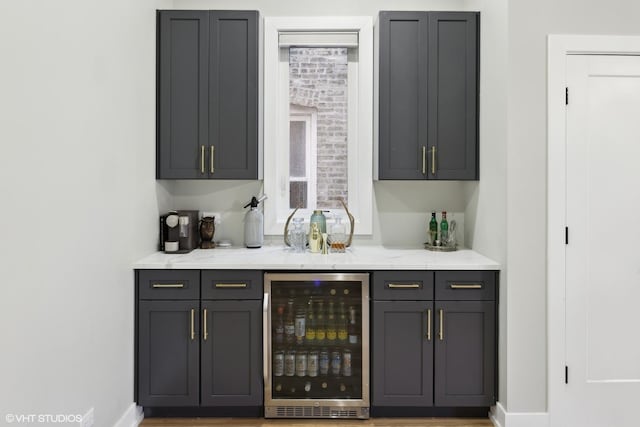 bar with wine cooler, light stone counters, and gray cabinets