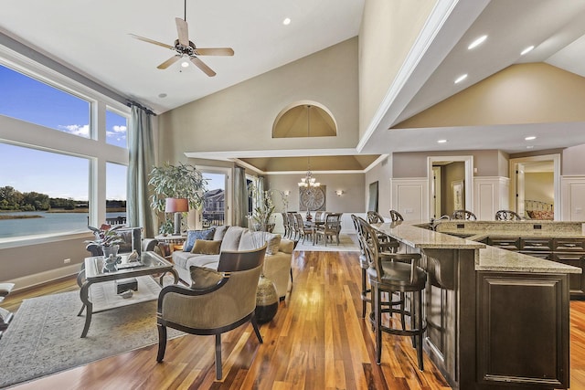 living room with sink, light hardwood / wood-style flooring, high vaulted ceiling, a water view, and ceiling fan with notable chandelier