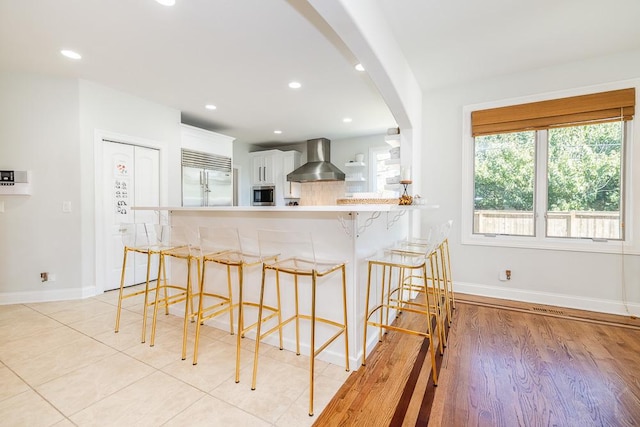 kitchen with a breakfast bar area, white cabinetry, built in appliances, kitchen peninsula, and wall chimney exhaust hood