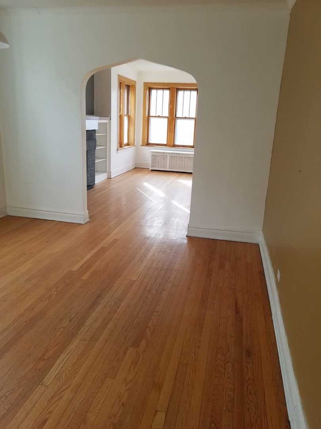 unfurnished living room featuring arched walkways, baseboards, radiator, light wood-style flooring, and a fireplace