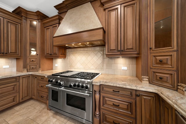 kitchen with range with two ovens, backsplash, custom exhaust hood, light stone countertops, and glass insert cabinets