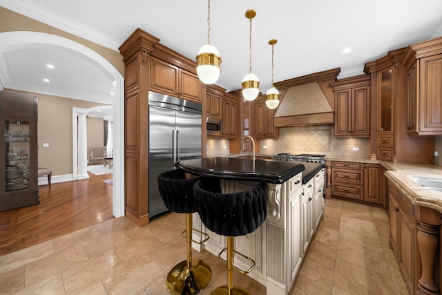 kitchen featuring arched walkways, brown cabinets, custom exhaust hood, and built in appliances