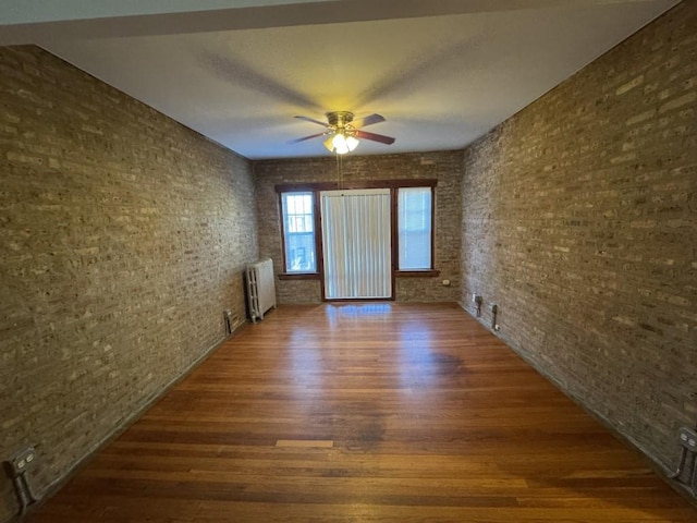 unfurnished room featuring brick wall, dark wood-type flooring, radiator heating unit, and ceiling fan