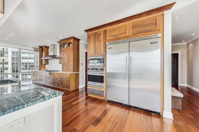 kitchen featuring brown cabinetry, wall chimney exhaust hood, built in appliances, crown molding, and a warming drawer