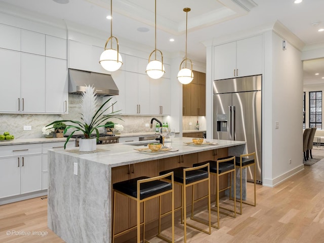 kitchen with white cabinetry, wall chimney range hood, an island with sink, and stainless steel built in fridge