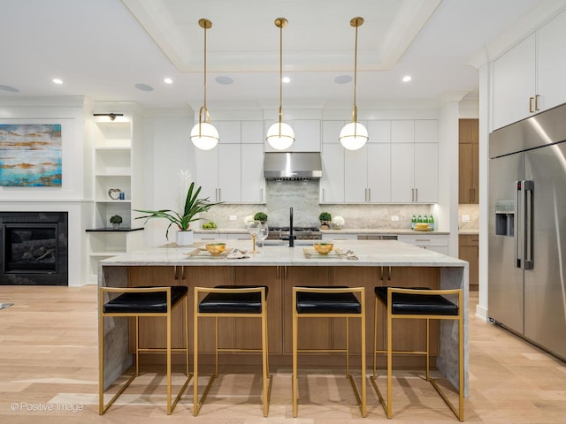 kitchen with extractor fan, white cabinetry, stainless steel built in refrigerator, a kitchen island with sink, and a tray ceiling