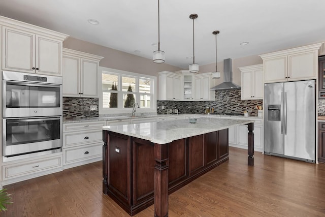 kitchen featuring a kitchen island, glass insert cabinets, appliances with stainless steel finishes, decorative light fixtures, and wall chimney range hood