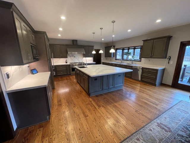 kitchen featuring pendant lighting, stainless steel appliances, dark wood-type flooring, a center island with sink, and wall chimney exhaust hood