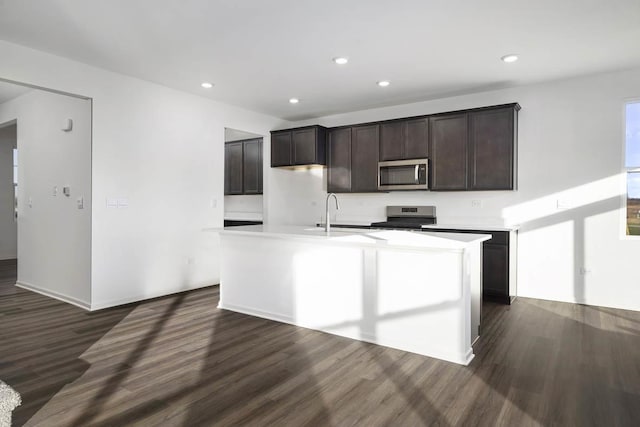 kitchen with sink, dark brown cabinets, stainless steel appliances, dark hardwood / wood-style floors, and a center island with sink