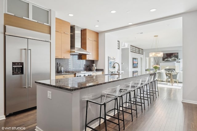 kitchen featuring wall chimney range hood, an island with sink, appliances with stainless steel finishes, and dark stone counters