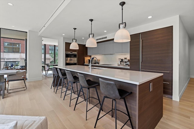 kitchen with sink, tasteful backsplash, light hardwood / wood-style flooring, hanging light fixtures, and a center island with sink