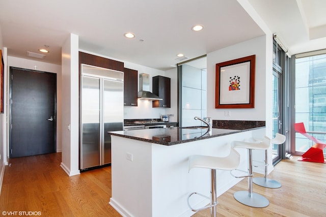 kitchen featuring expansive windows, a kitchen bar, built in fridge, wall chimney exhaust hood, and light wood-type flooring