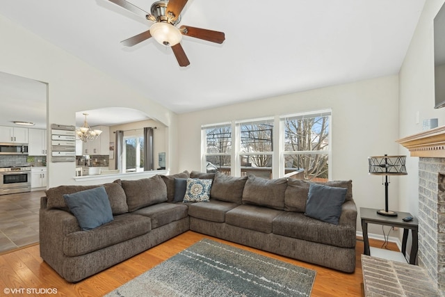 living room featuring ceiling fan with notable chandelier, light hardwood / wood-style floors, vaulted ceiling, and a brick fireplace