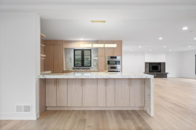 kitchen with light brown cabinets, kitchen peninsula, double oven, light stone countertops, and light hardwood / wood-style floors