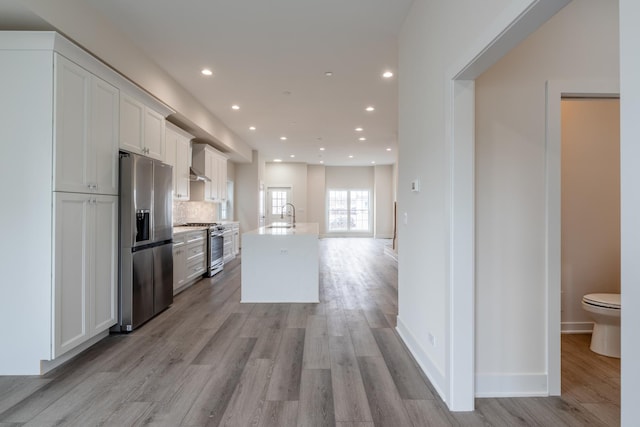 kitchen featuring appliances with stainless steel finishes, tasteful backsplash, white cabinets, a center island with sink, and light wood-type flooring