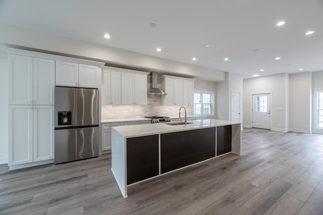 kitchen with sink, white cabinetry, stainless steel refrigerator with ice dispenser, a center island with sink, and wall chimney exhaust hood