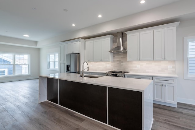 kitchen with wall chimney range hood, sink, stainless steel fridge, white cabinetry, and a center island with sink