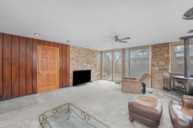 carpeted living room with a wall of windows, wooden walls, ceiling fan, and a wood stove