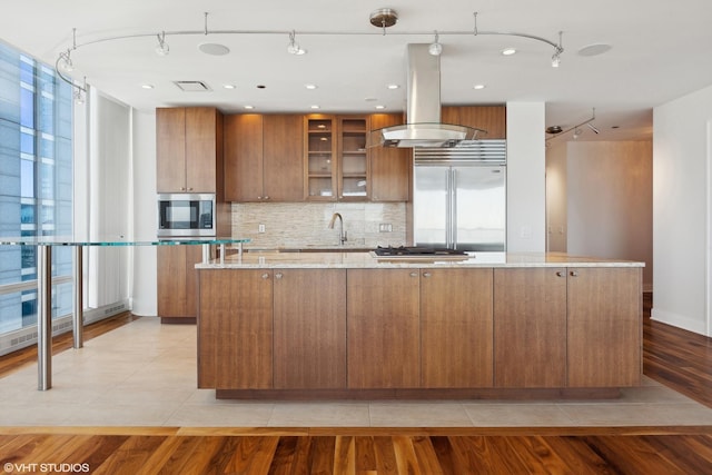 kitchen with island range hood, decorative backsplash, a large island, brown cabinets, and stainless steel appliances