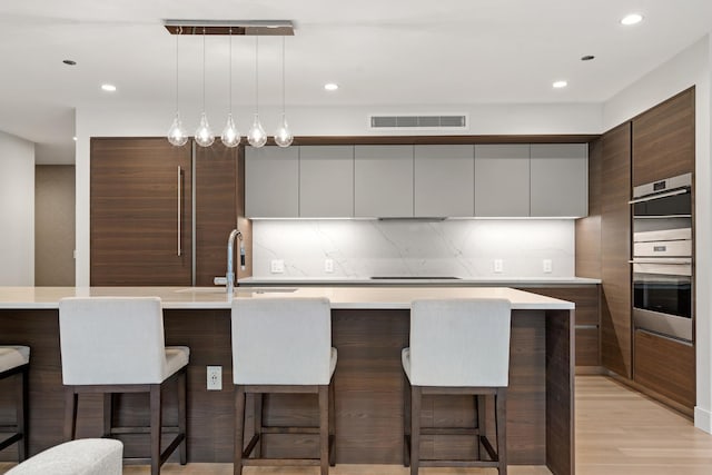 kitchen featuring sink, decorative backsplash, a center island with sink, decorative light fixtures, and stainless steel double oven