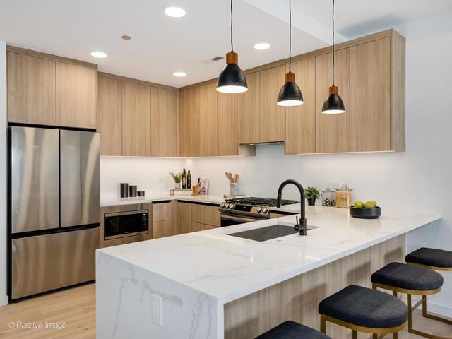 kitchen with appliances with stainless steel finishes, hanging light fixtures, light stone counters, light brown cabinetry, and kitchen peninsula