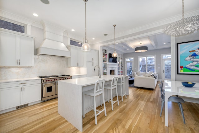 kitchen with premium range hood, white cabinetry, double oven range, a kitchen island with sink, and decorative light fixtures