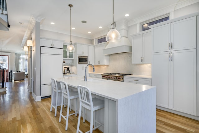 kitchen with crown molding, white cabinetry, built in appliances, a center island with sink, and decorative light fixtures