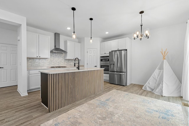 kitchen featuring wall chimney range hood, a kitchen island with sink, white cabinetry, hanging light fixtures, and stainless steel appliances