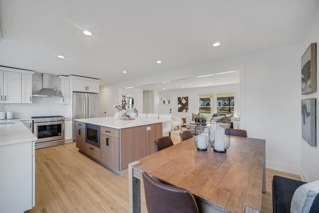 kitchen with appliances with stainless steel finishes, a kitchen island, wall chimney range hood, light hardwood / wood-style floors, and white cabinets