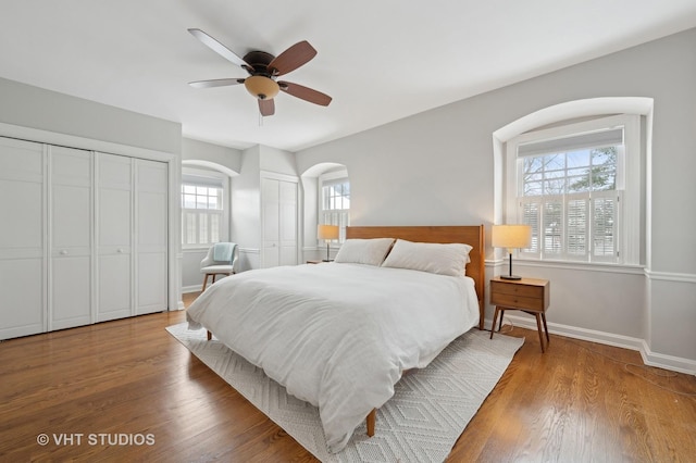 bedroom featuring a ceiling fan, wood finished floors, baseboards, arched walkways, and multiple closets