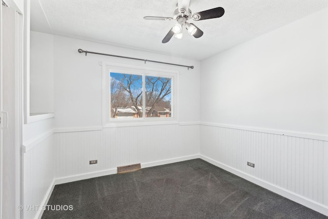 spare room with a textured ceiling, visible vents, a ceiling fan, wainscoting, and dark colored carpet