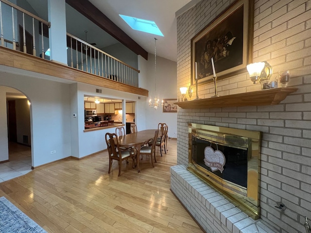 dining space featuring high vaulted ceiling, a skylight, a notable chandelier, a brick fireplace, and light hardwood / wood-style flooring