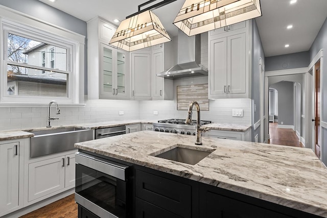 kitchen featuring sink, light stone counters, decorative light fixtures, stainless steel appliances, and wall chimney range hood