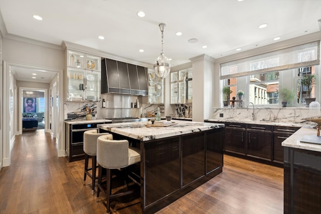 kitchen featuring light stone counters, a kitchen bar, glass insert cabinets, a kitchen island, and wall chimney exhaust hood