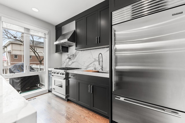 kitchen with wall chimney range hood, high end appliances, dark cabinets, and light wood finished floors