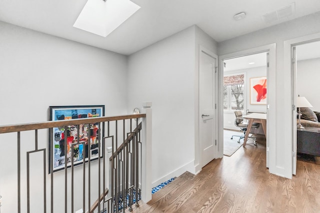 corridor featuring a skylight, wood finished floors, an upstairs landing, visible vents, and baseboards