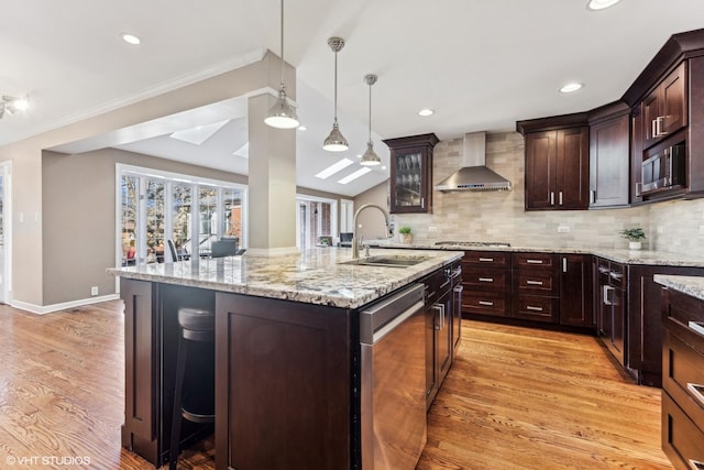 kitchen with glass insert cabinets, a kitchen island with sink, wall chimney range hood, pendant lighting, and a sink