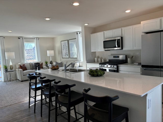 kitchen with a kitchen island with sink, a sink, white cabinets, open floor plan, and appliances with stainless steel finishes