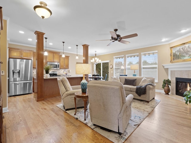 living room with crown molding, light hardwood / wood-style flooring, decorative columns, a tiled fireplace, and ceiling fan with notable chandelier