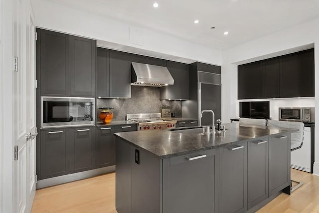 kitchen with stainless steel microwave, an island with sink, sink, light wood-type flooring, and wall chimney exhaust hood