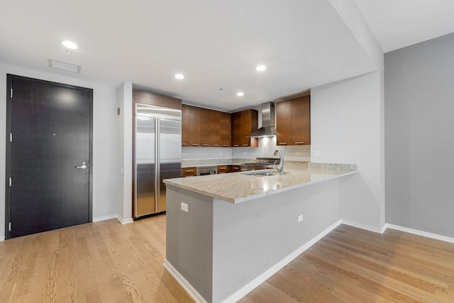 kitchen with a sink, wall chimney range hood, light wood-type flooring, stainless steel built in refrigerator, and a peninsula