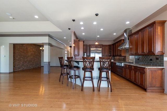 kitchen featuring a breakfast bar area, appliances with stainless steel finishes, light wood-type flooring, wall chimney exhaust hood, and decorative columns